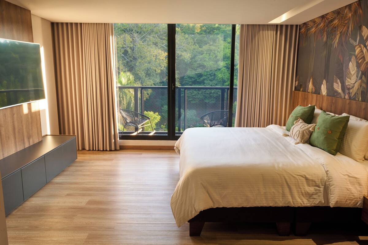 A modern bedroom with a large bed, a TV mounted on the wall, and a balcony with a view of trees.