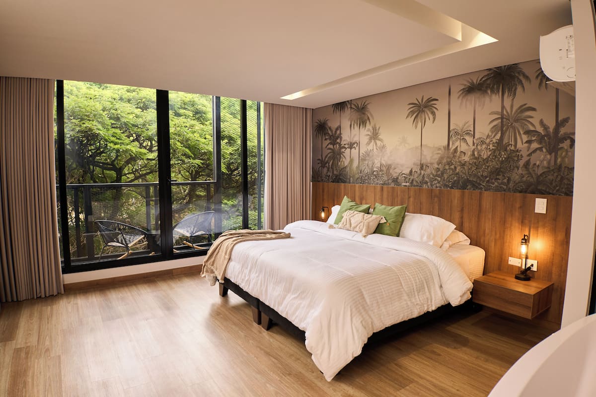 A bedroom with a large window, a bed, a nightstand, and a wall mural.
