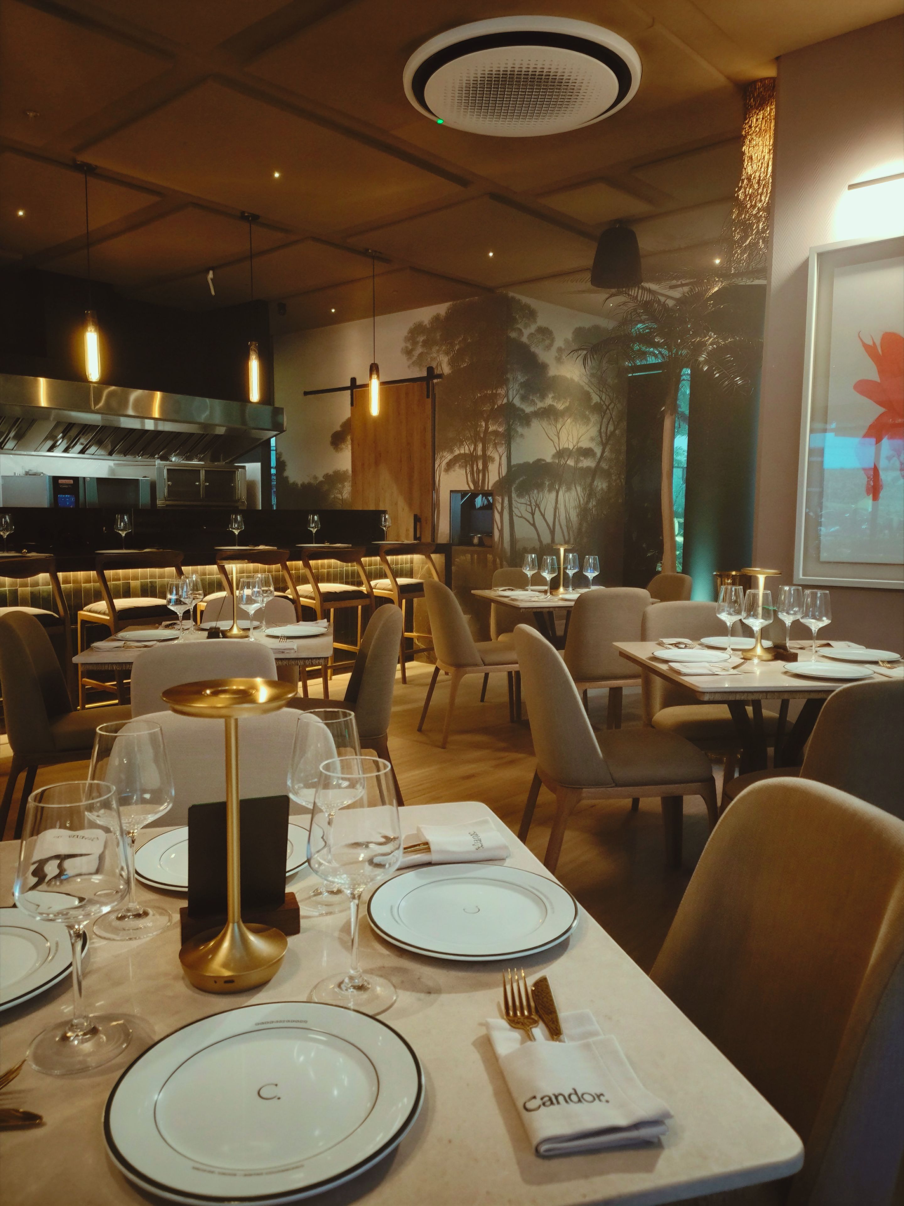 A restaurant interior with multiple dining tables, chairs, and a kitchen area in the background.