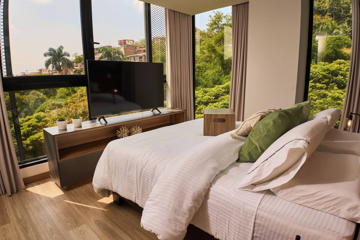 A modern bedroom with a large window, a bed, a TV on a stand, and a small table.
