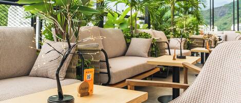 An outdoor seating area with tables and chairs, surrounded by potted plants and a glass ceiling.