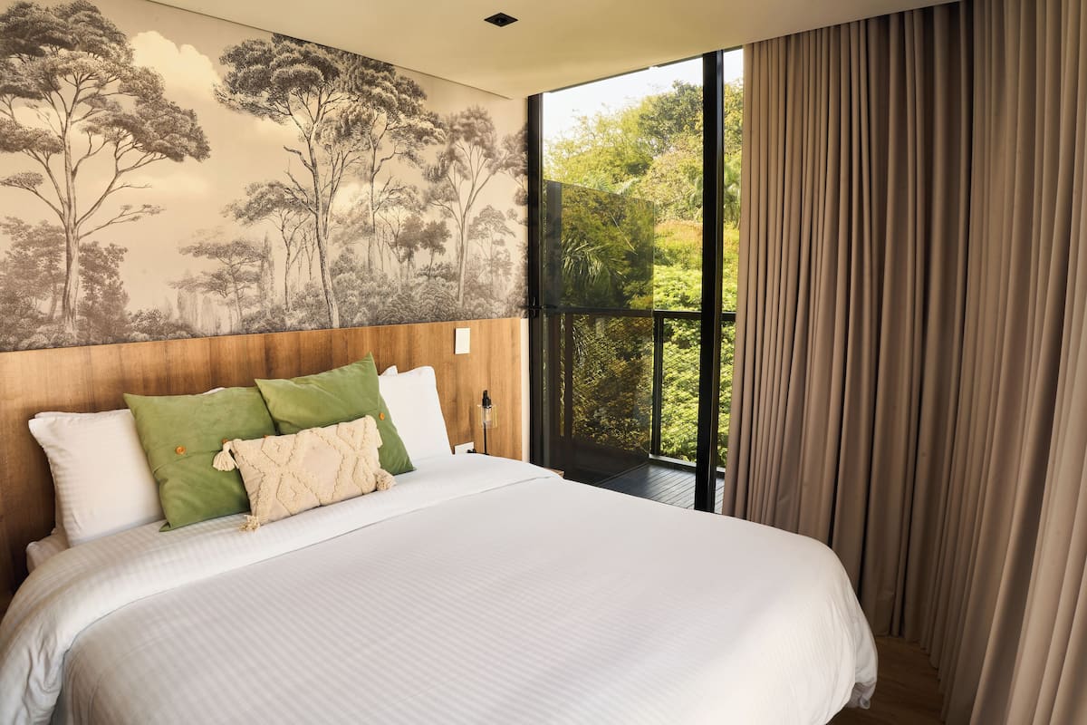 A bedroom with a large bed, a wooden headboard, a mural of trees, and a view through a window with curtains.