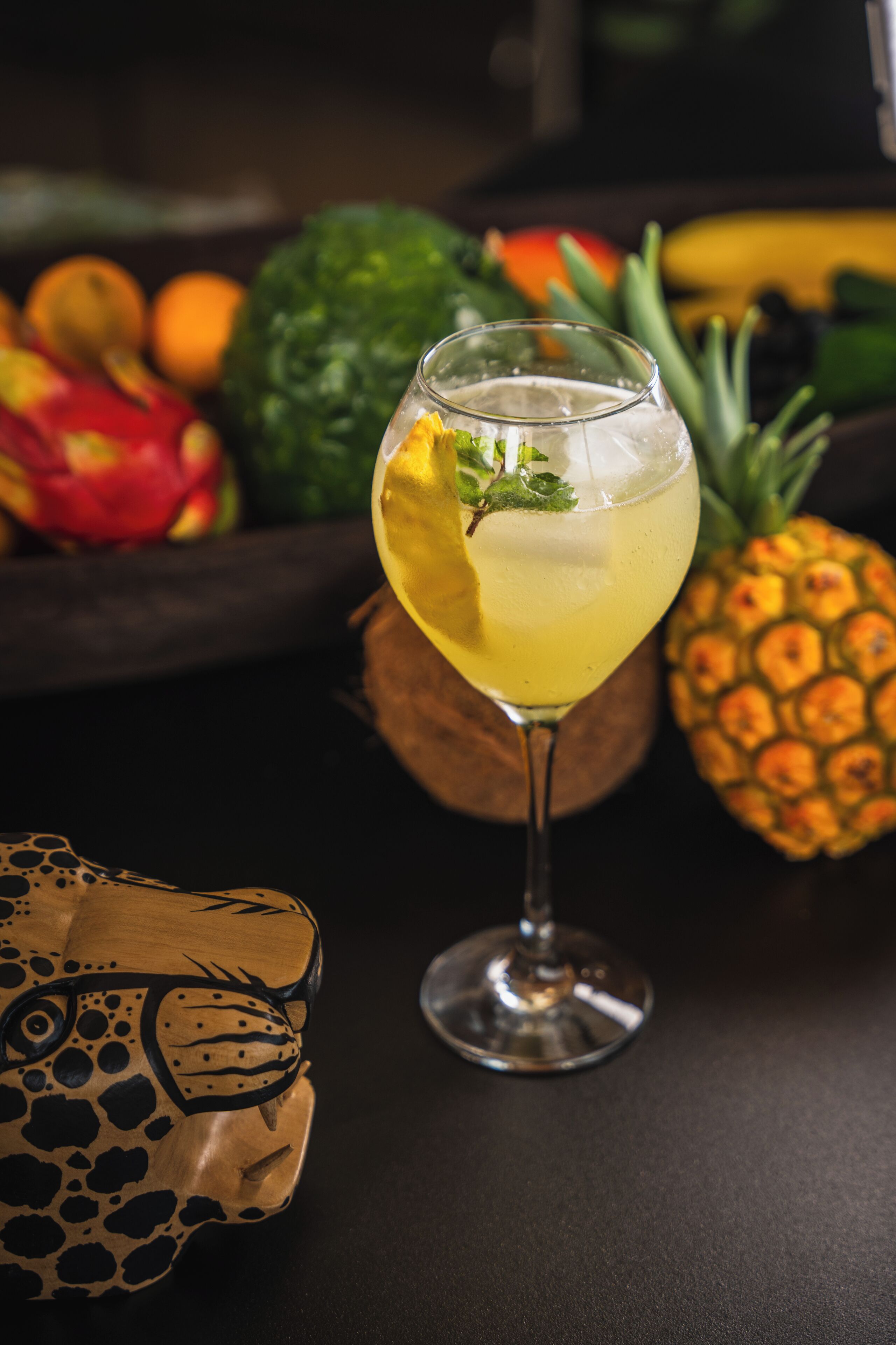 A glass of yellow beverage with a garnish, a pineapple, and a bowl of fruit in the background.