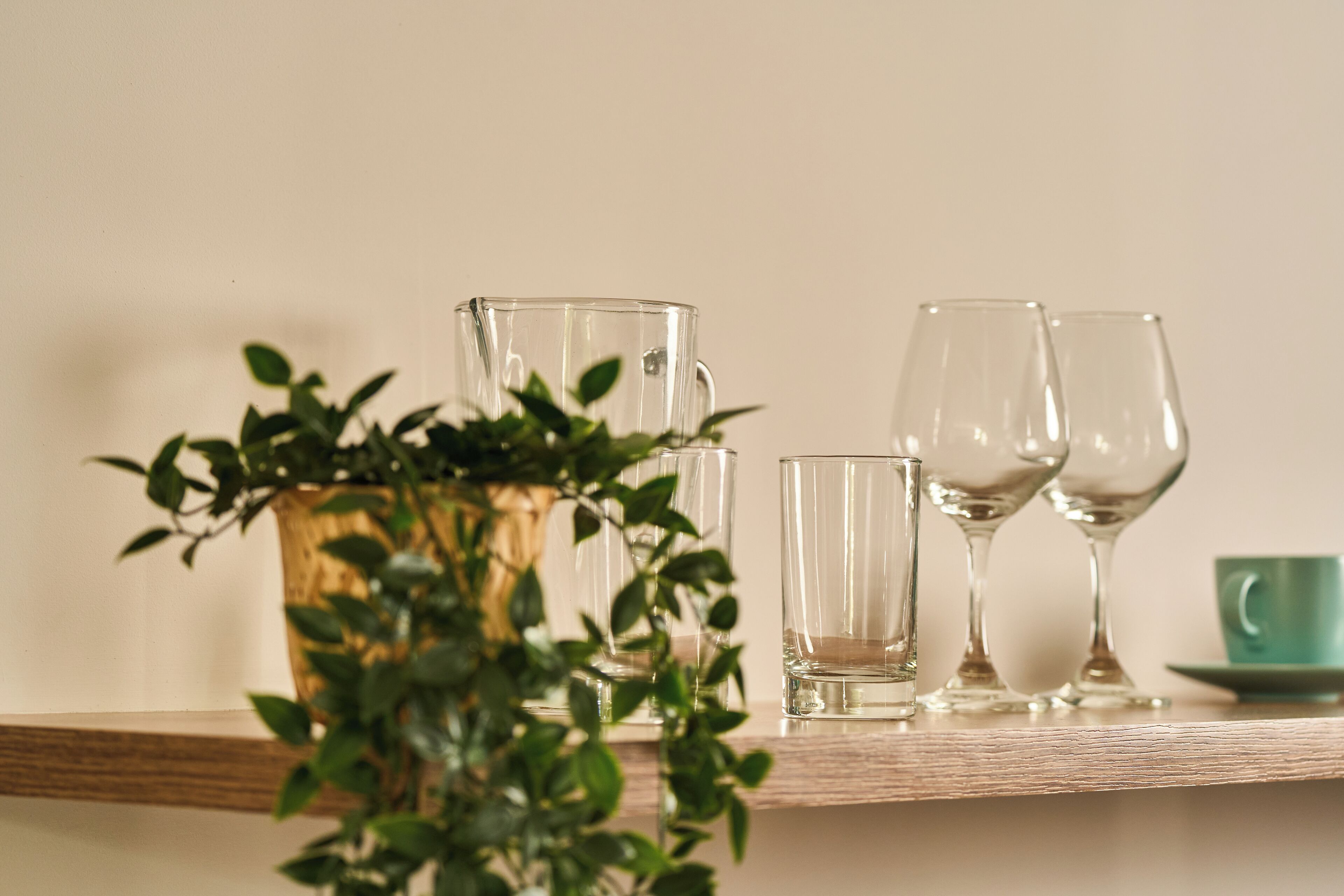 A wooden shelf with a clear glass pitcher, two wine glasses, and a green cup.