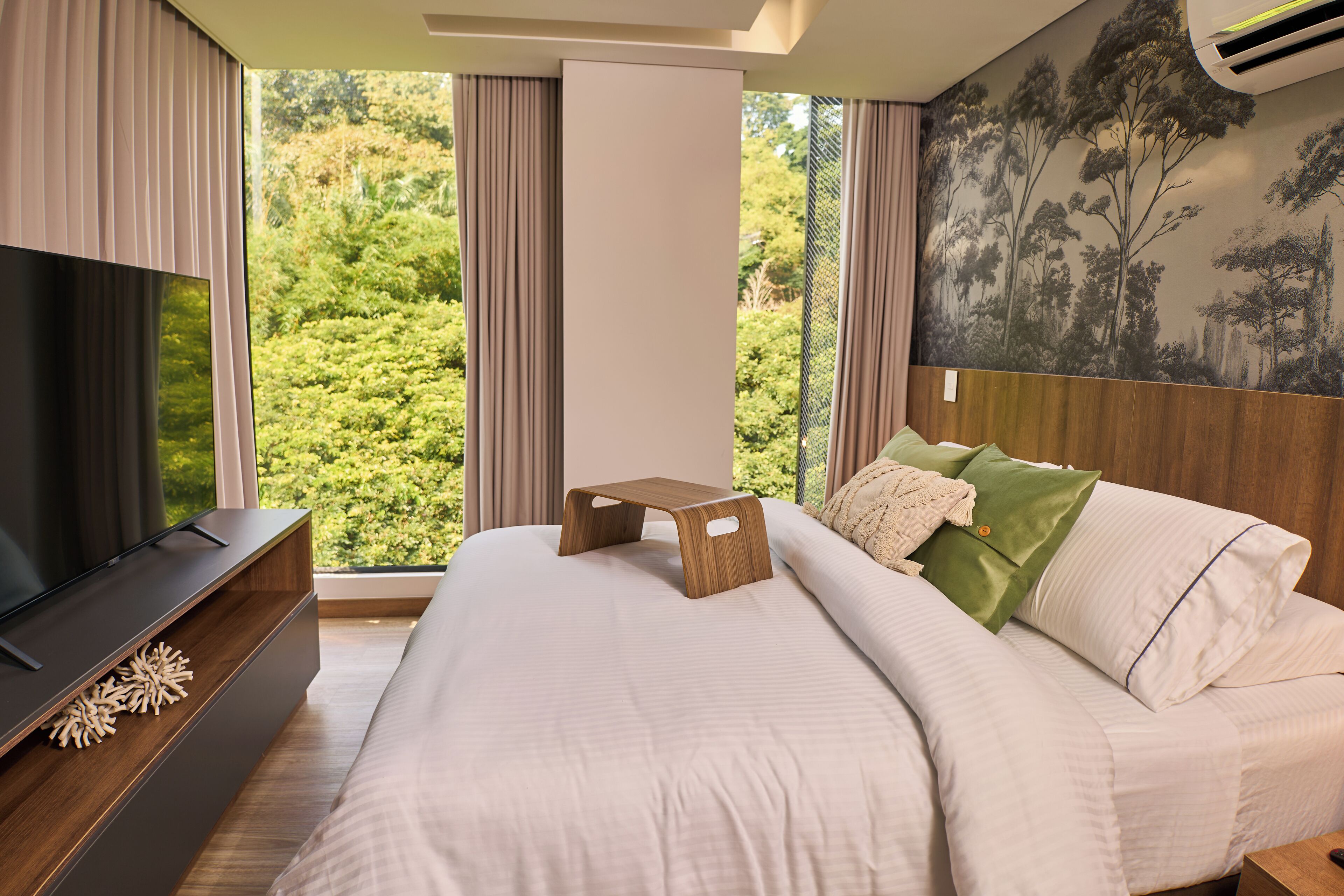 A bedroom with a bed, a TV, and a view of greenery through large windows.