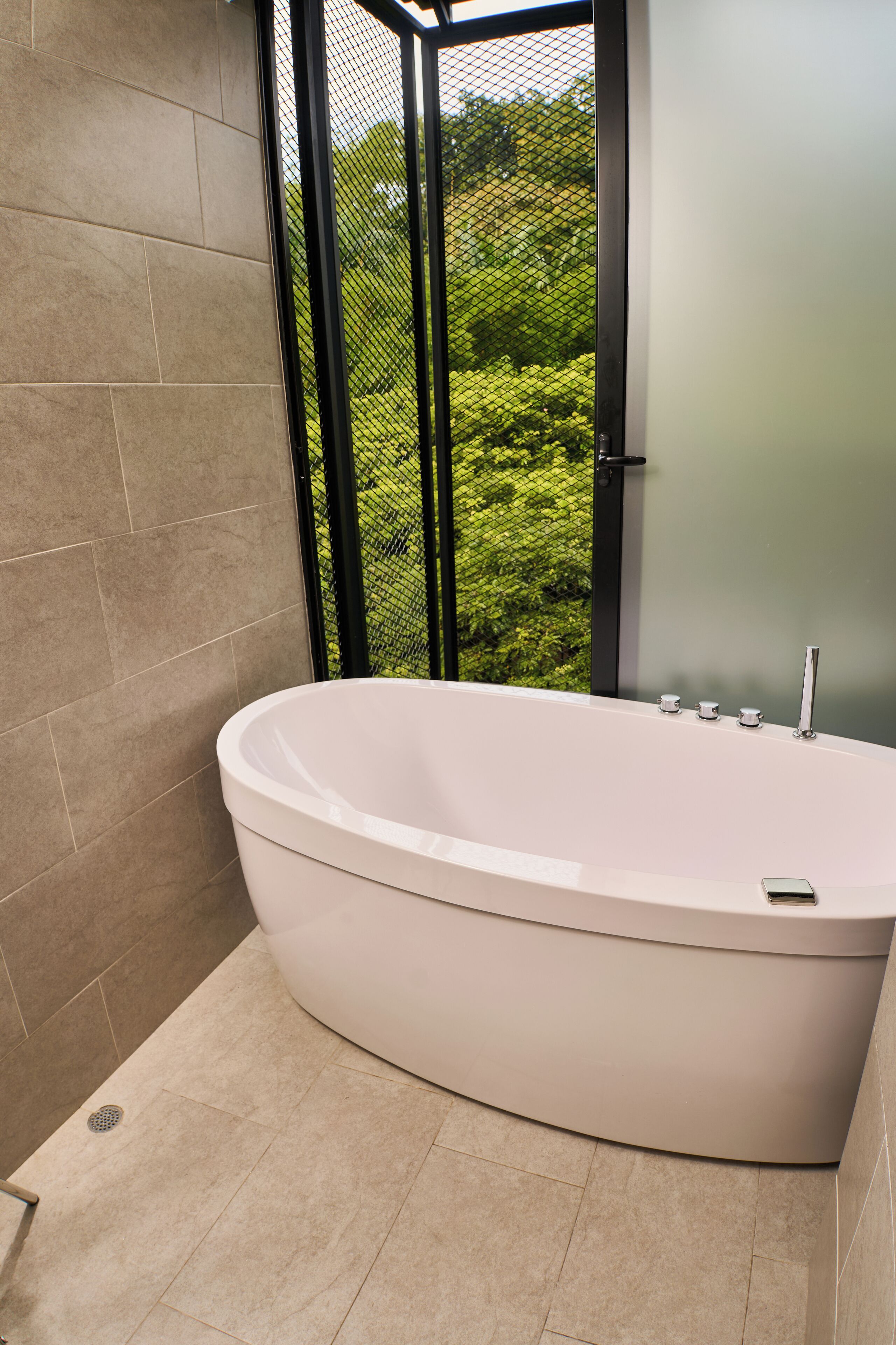 A modern bathroom with a freestanding bathtub, a window with a view of greenery, and a tiled floor.