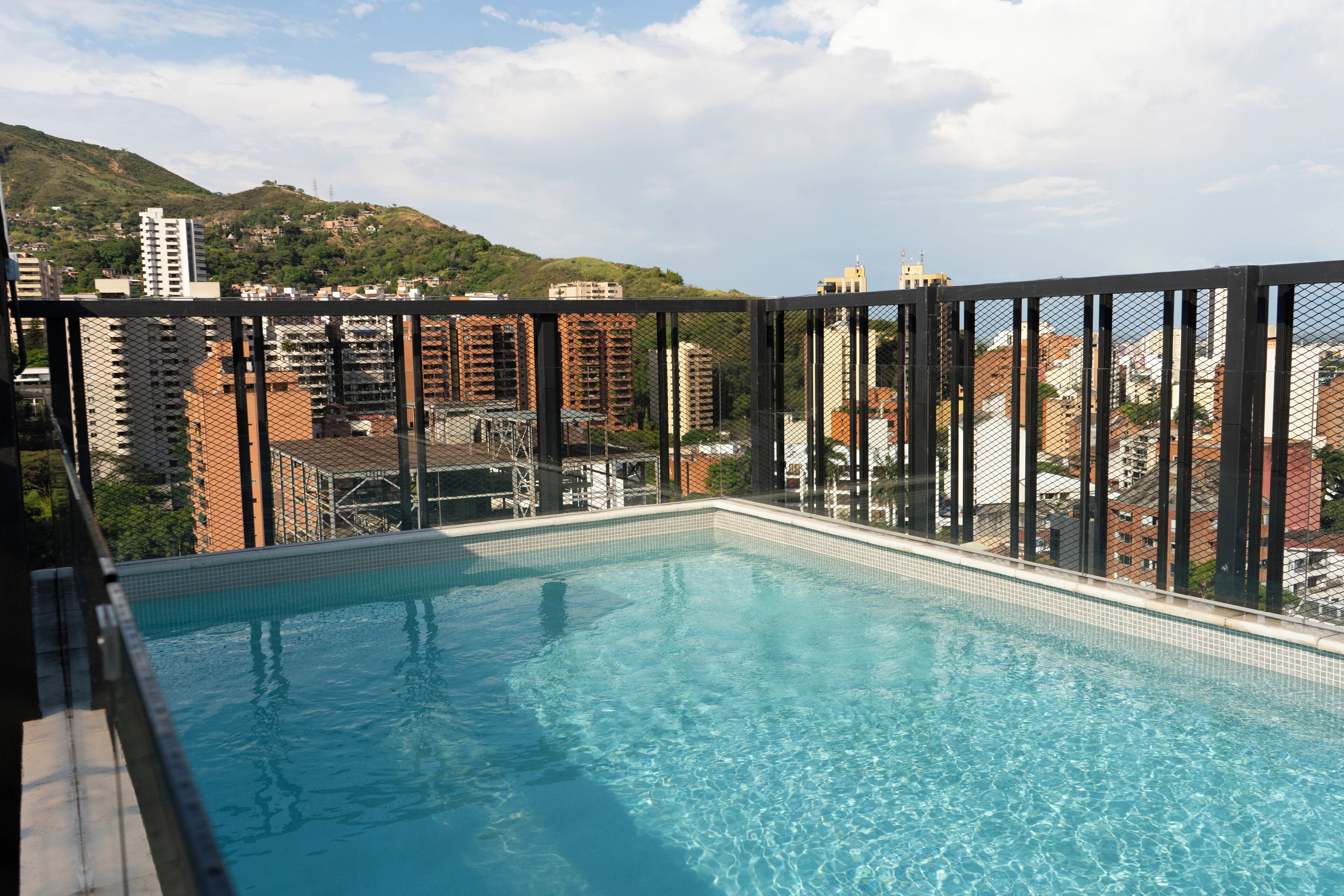 Una piscina rectangular con borde de azulejos, rodeada por una cerca de metal, con edificios urbanos y un paisaje montañoso de fondo.