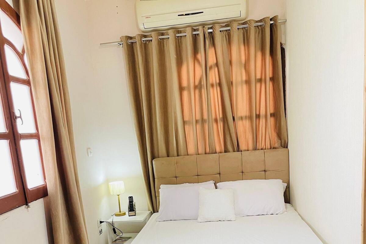 A bedroom with a bed, curtains, an air conditioning unit, a nightstand, and a lamp.