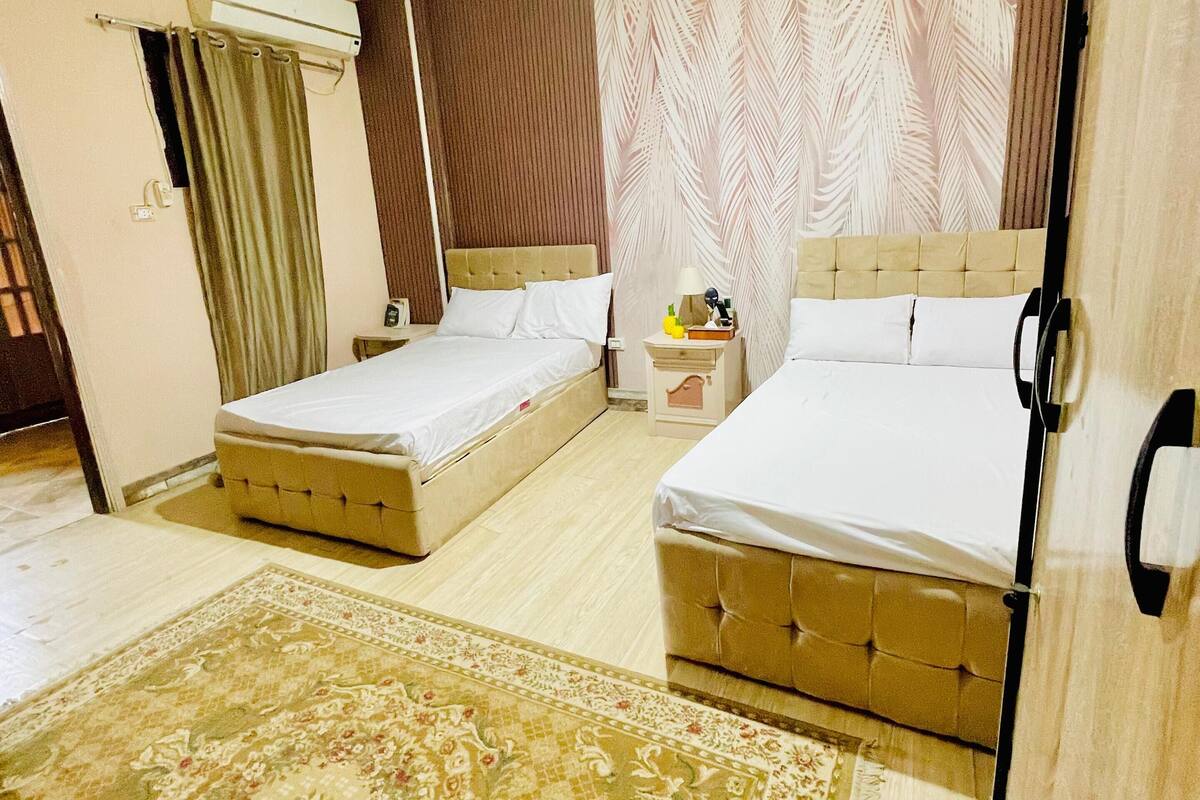 A double room with two beds, a large decorative rug, a ceiling light, and a window with curtains.