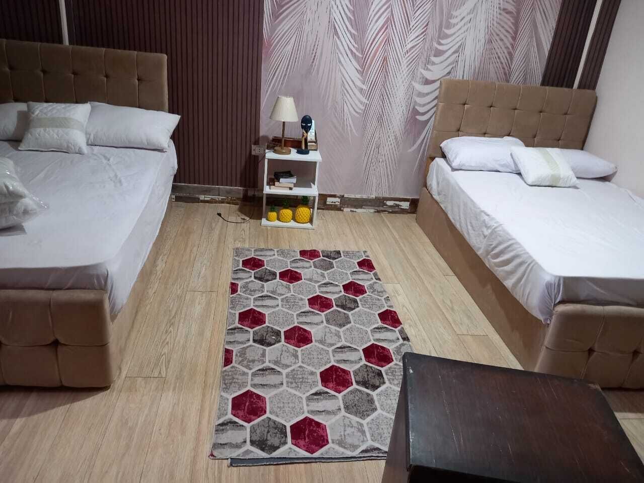 A room with two beds, a small shelf with books and decorative items, and a patterned rug.