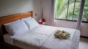 Ein ordentlich bezogenes Bett mit weißer Decke und Kissen, ein Holz-Kopfbrett und ein Fenster mit Blick ins Grüne.