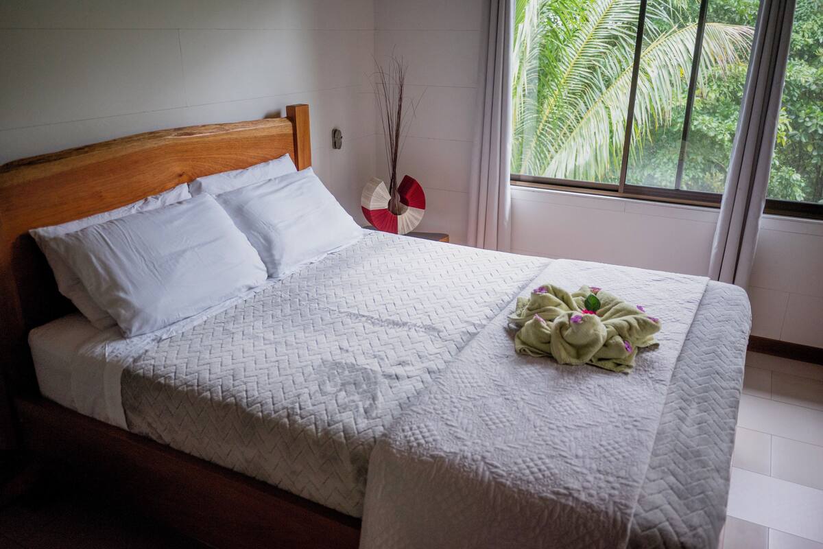A neatly made bed with a white quilt and pillows, a wooden headboard, and a window with a view of greenery.