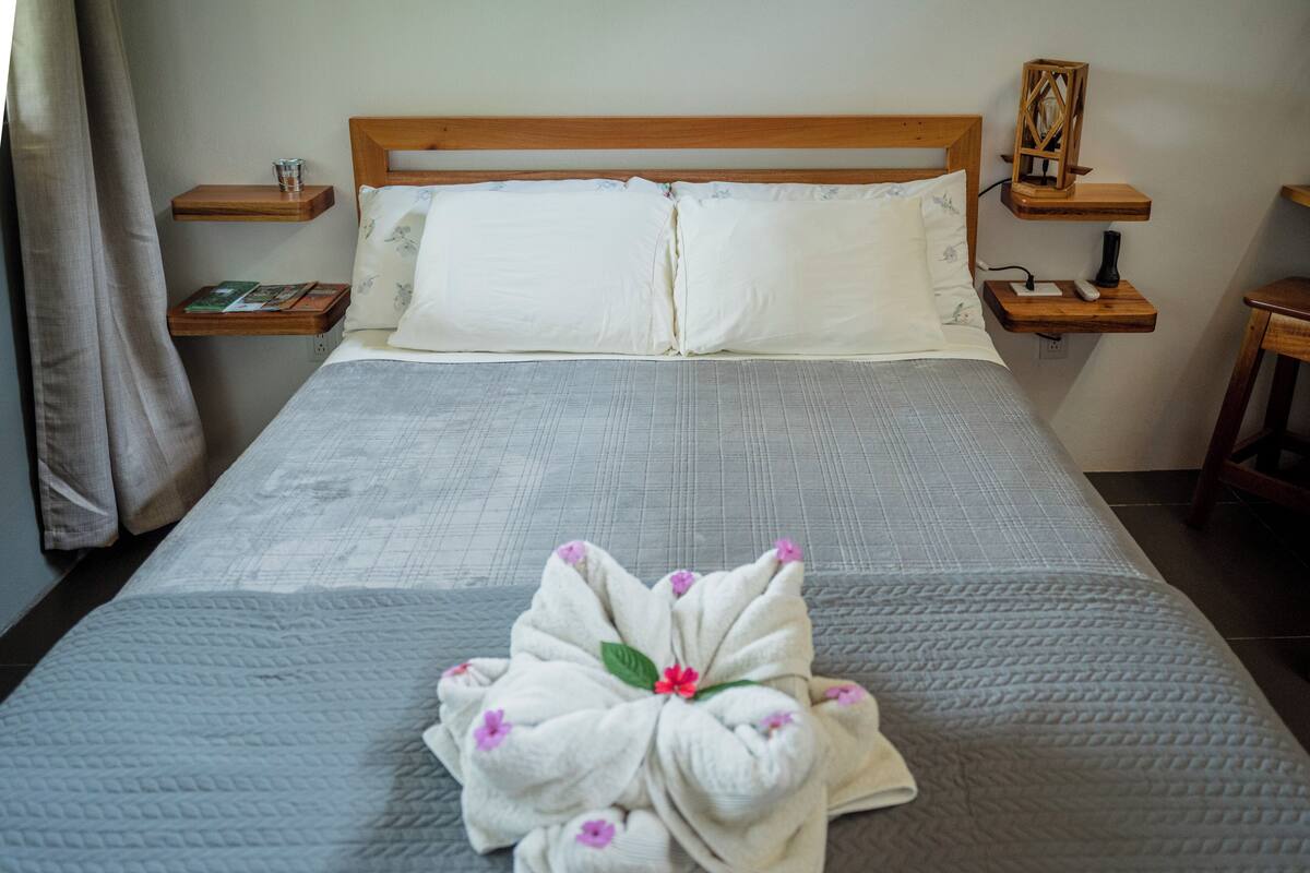 A neatly made bed with a white comforter, two pillows, and a floral towel on the bed. There are wooden nightstands with books and a lamp on them.