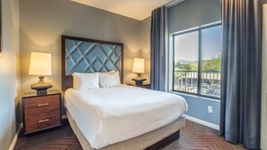 A hotel room with a large bed, two bedside tables with lamps, a view of a balcony, and a large window with curtains.