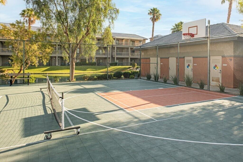 An outdoor basketball court with a fence, trees, and a building in the background.