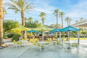 An outdoor patio with white tables and chairs, green and blue umbrellas, and a barbecue area.