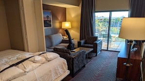 A hotel room with a bed, a sofa, a desk, and a chair. There is a window with a view of palm trees and buildings. - Indio | Desert Couples Getaway | Pool & Lazy River (Indio)