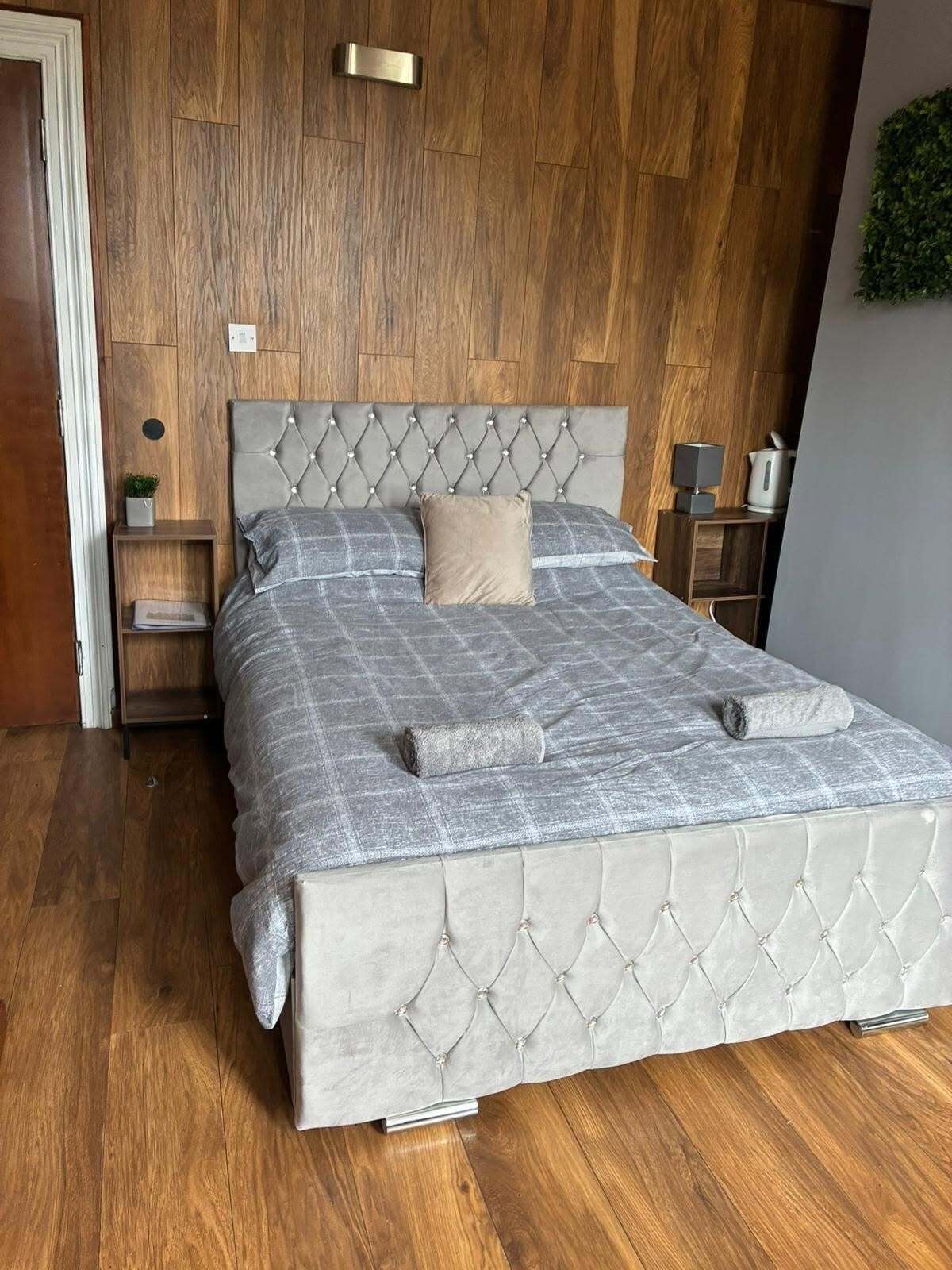 Un lit avec une tête de lit capitonnée, une table de chevet, une petite étagère et un luminaire fixé au mur.