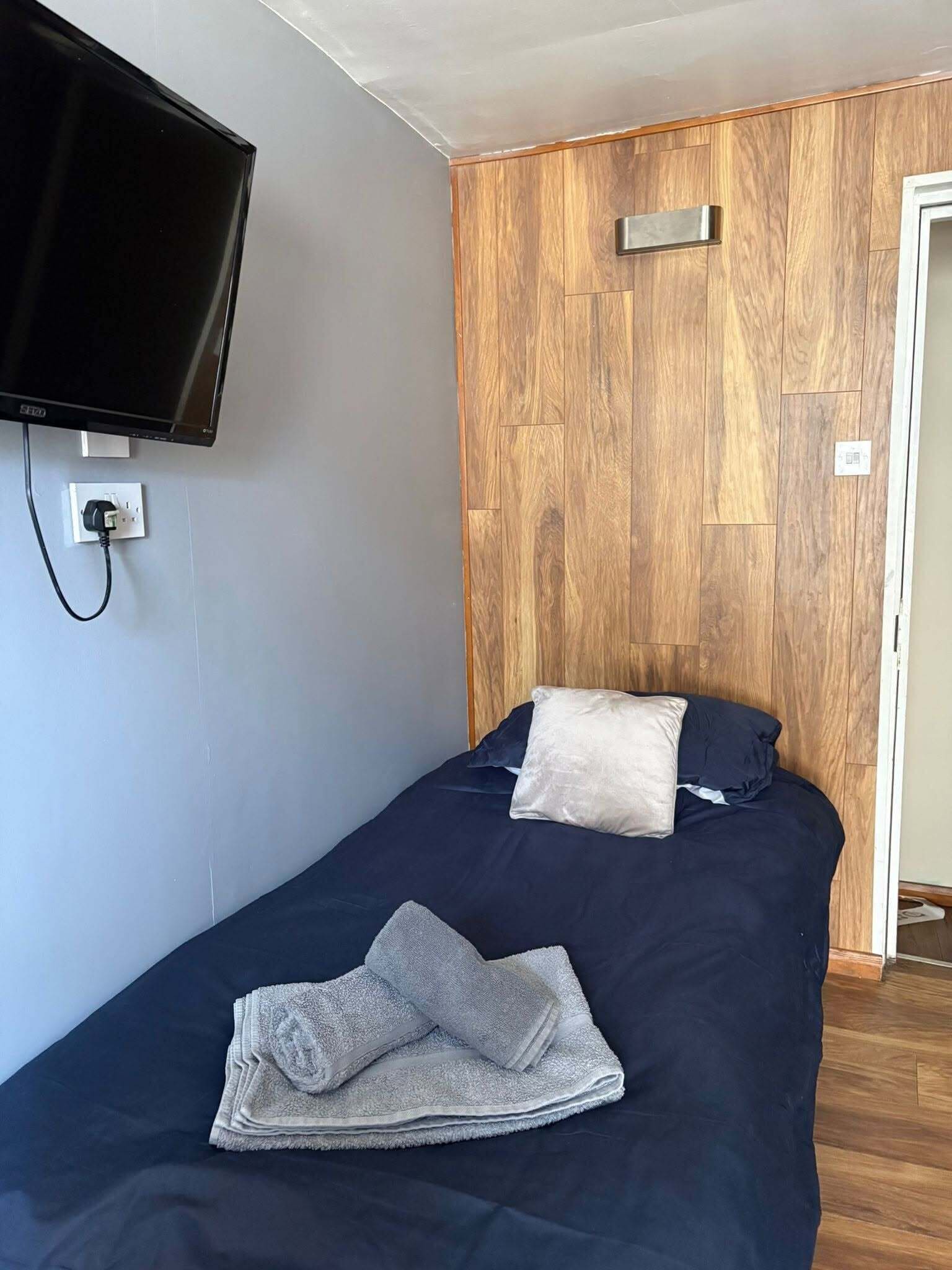 A single bed with a dark blue bedspread, two pillows, a folded towel, a mounted television, and a wooden panel wall.