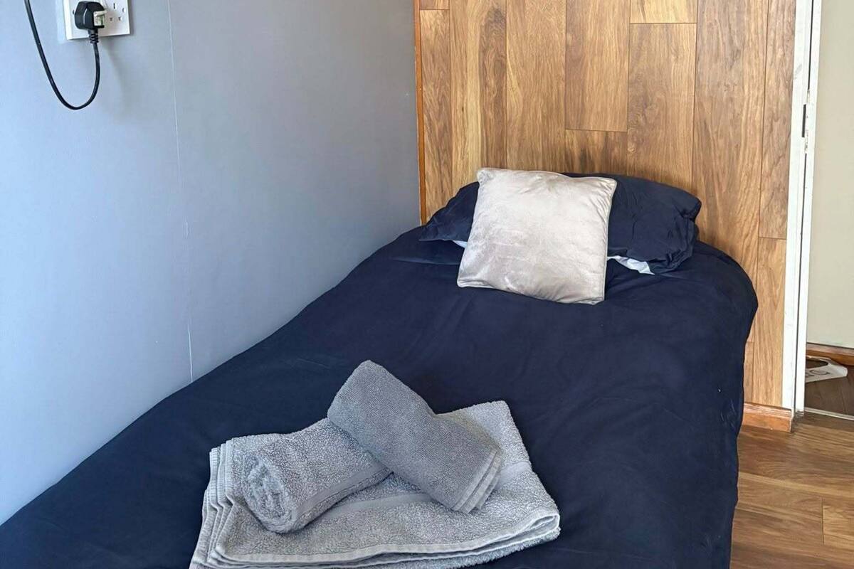 Een enkel bed met een donkerblauwe spreideken, twee kussens, een opgevouwen handdoek, een bevestigde televisie en een houten paneelwand.