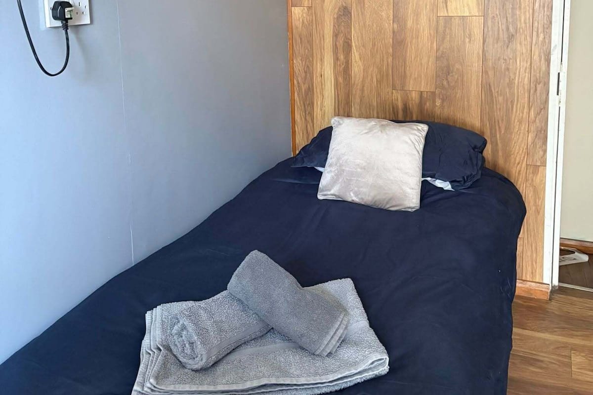 A single bed with a dark blue bedspread, two pillows, a folded towel, a mounted television, and a wooden panel wall.