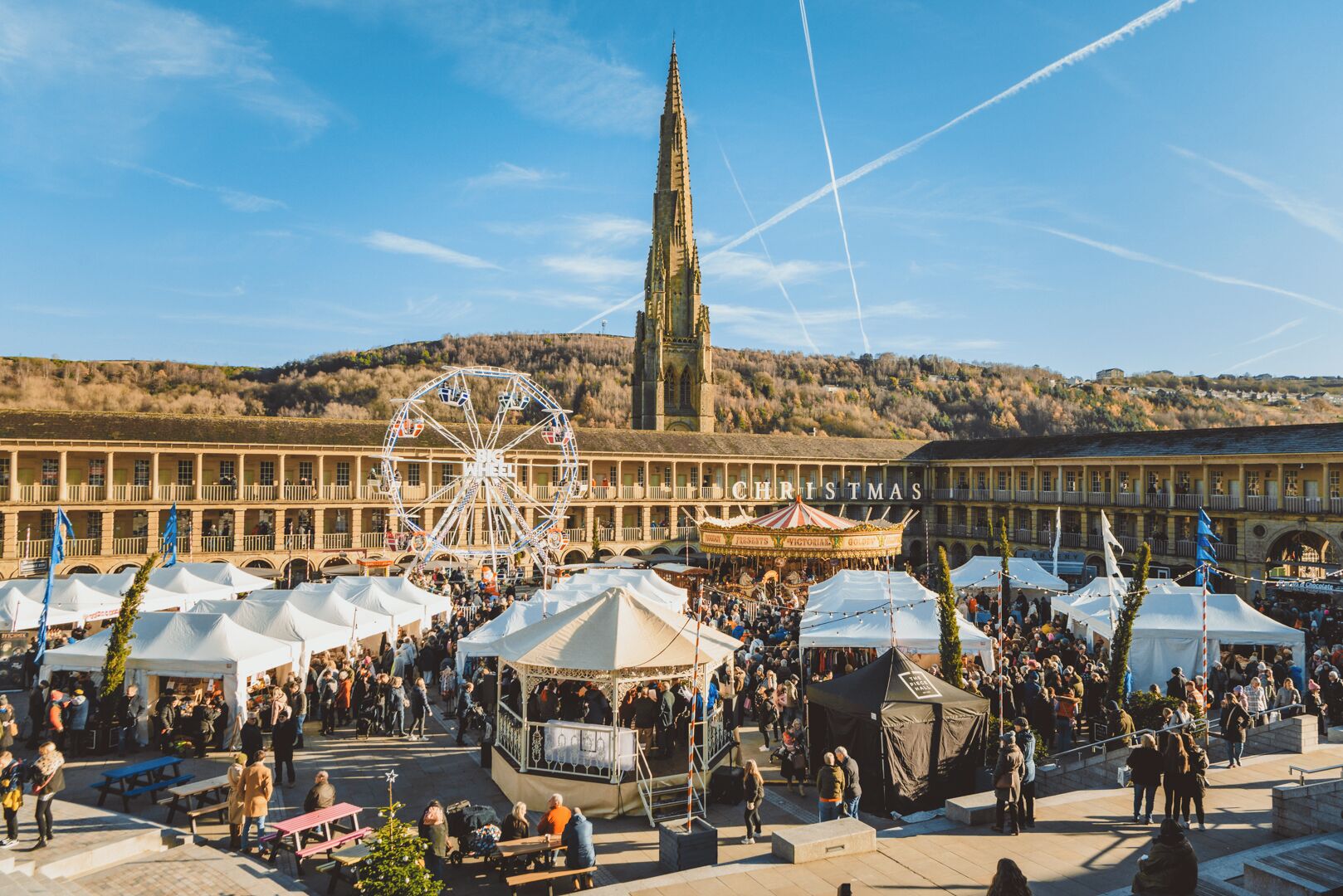 A historical building with a prominent tower, a large Ferris wheel, and a festive market area with tents and people.