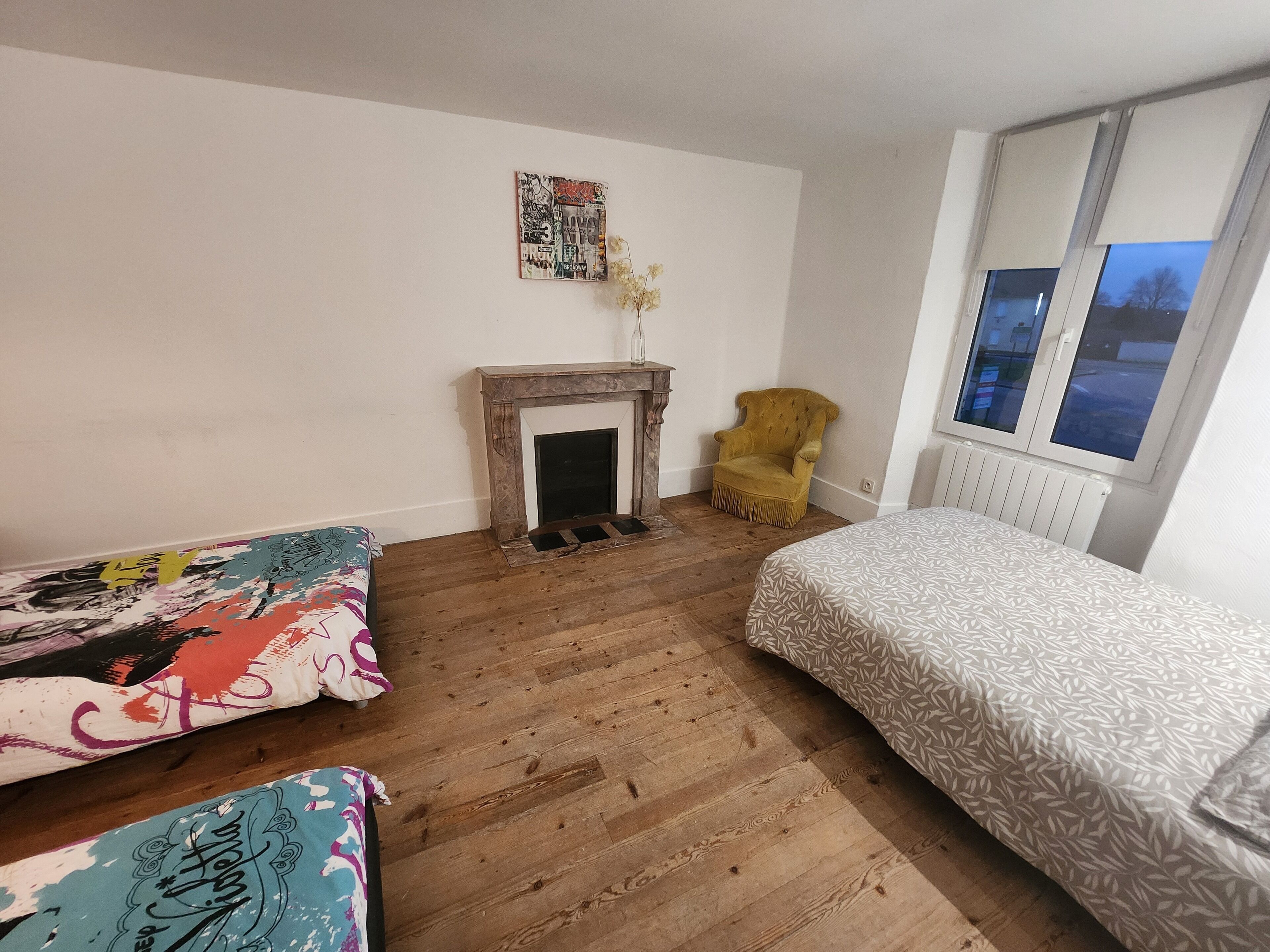 A bedroom with a wooden floor, a fireplace, a yellow armchair, and a bed with patterned bedding.