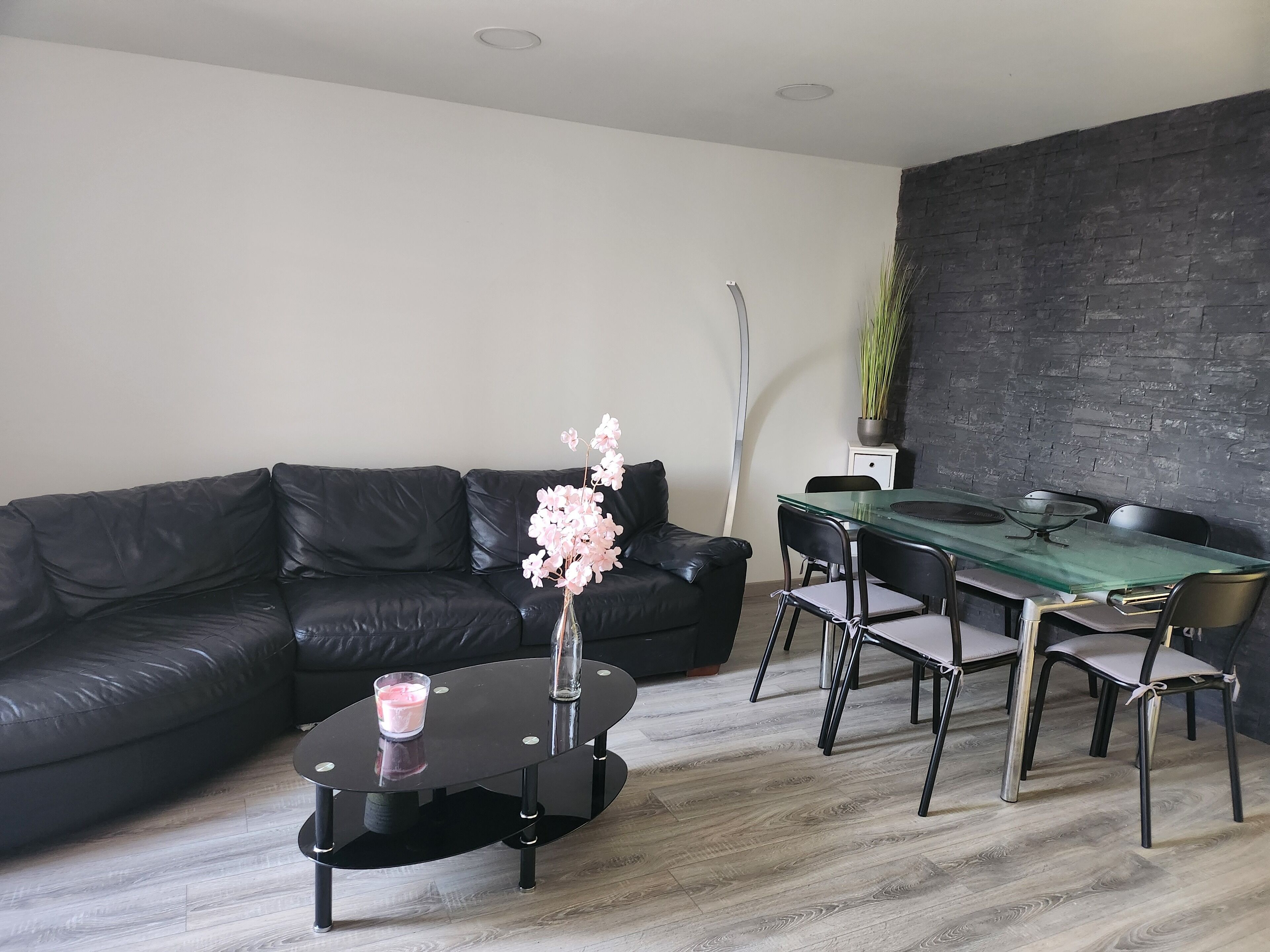A modern living room with a black leather sofa, a glass-top dining table with chairs, a small black coffee table with a vase of flowers, and a tall floor lamp.