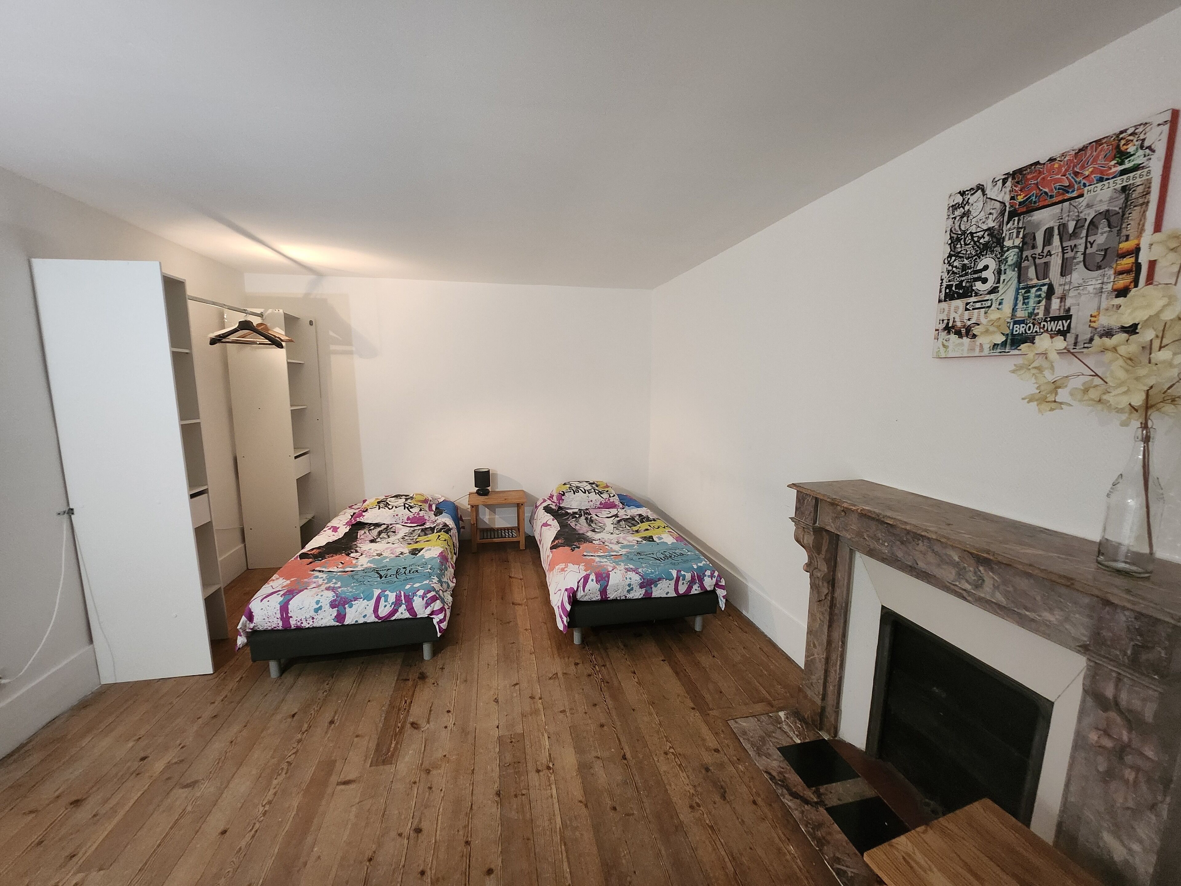 A bedroom with two beds, a wooden floor, a wardrobe, a small table, and a fireplace.