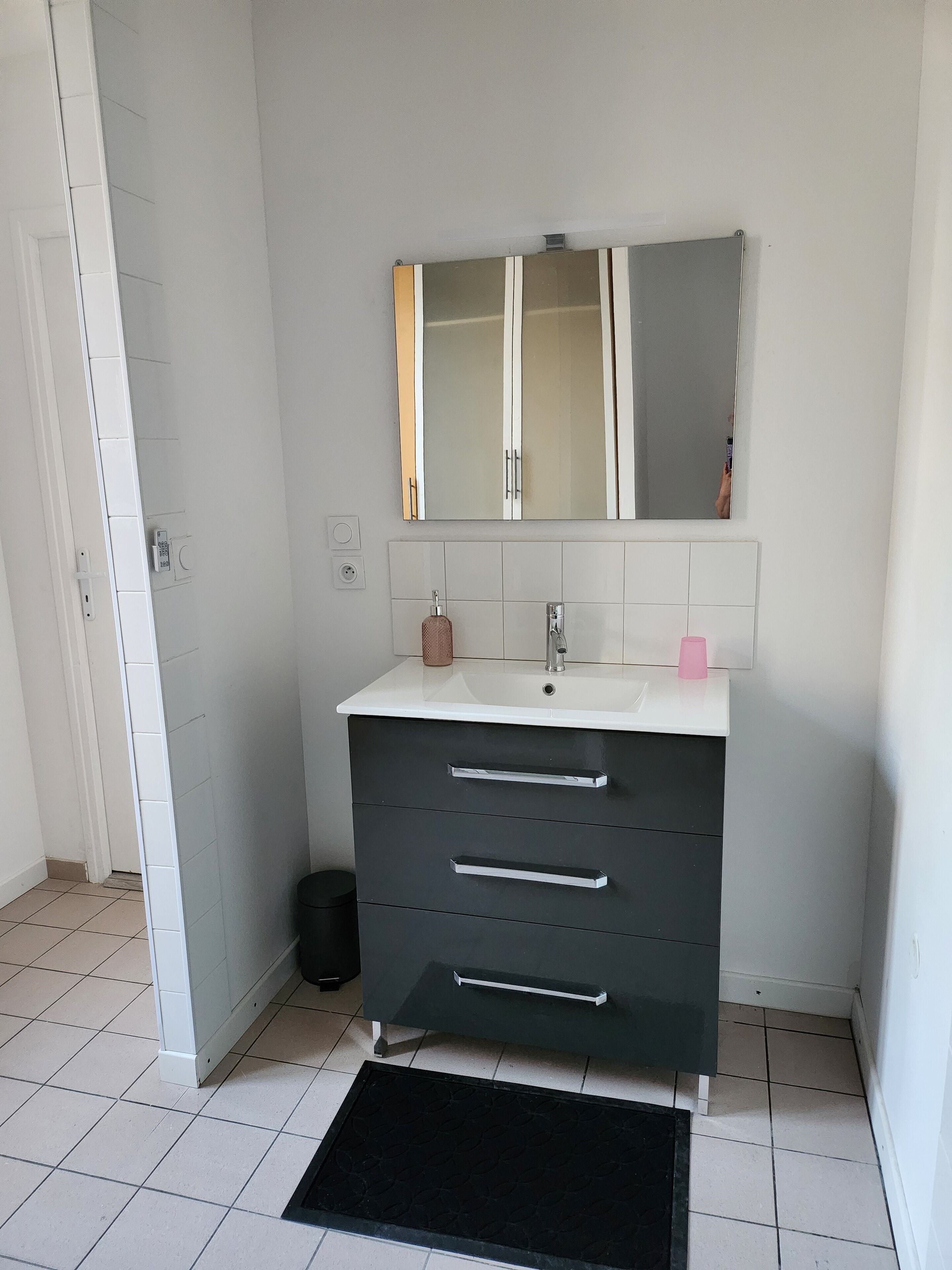 Un baño con un lavamanos blanco, un espejo y un tapete negro en el suelo.