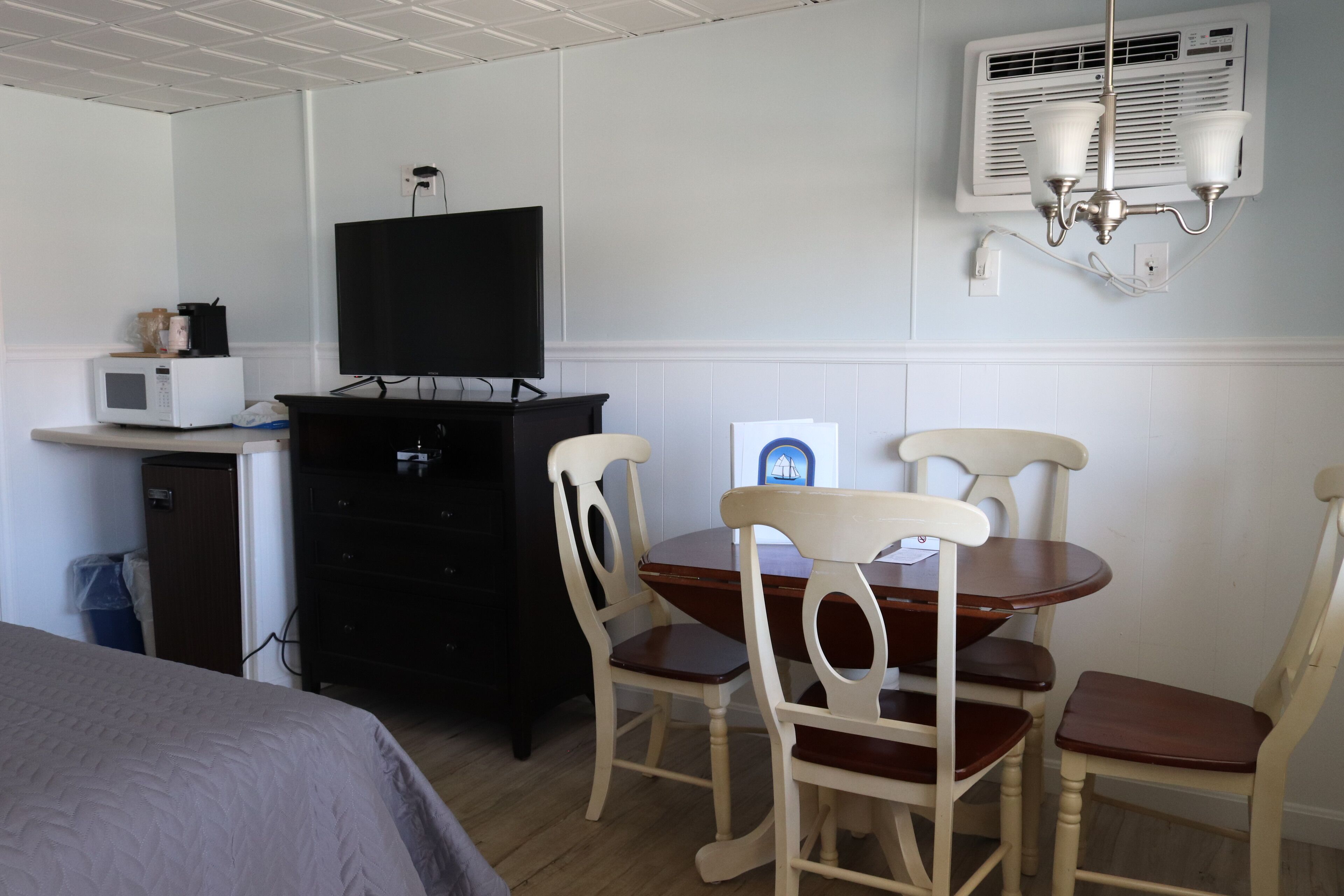 Una habitación con una cama, un televisor sobre un aparador, un microondas, un mueble pequeño, una mesa de comedor con sillas y un aire acondicionado instalado en la pared.