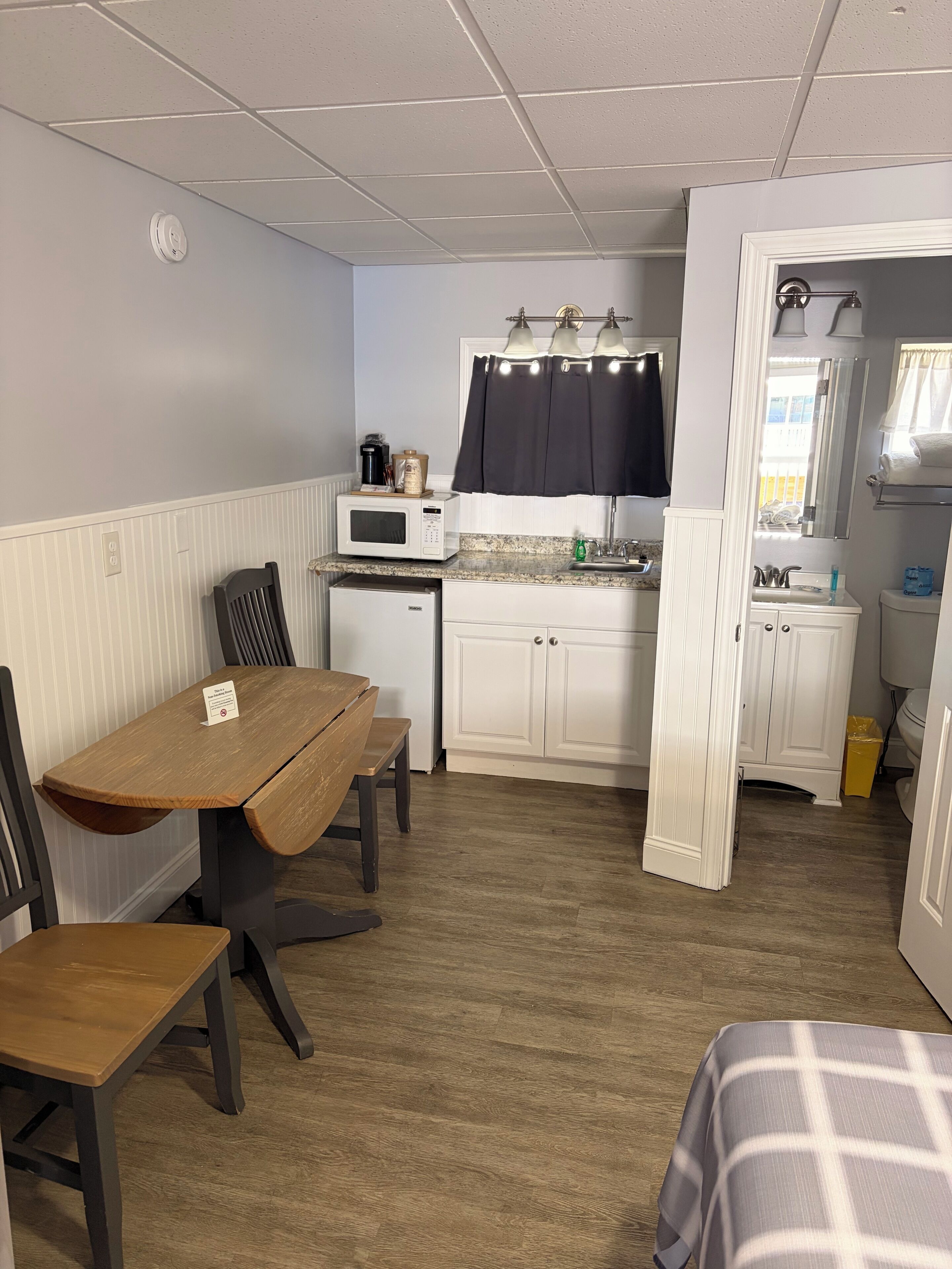 A compact kitchen with a microwave, sink, and cabinet, a dining table with chairs, and a small bench.