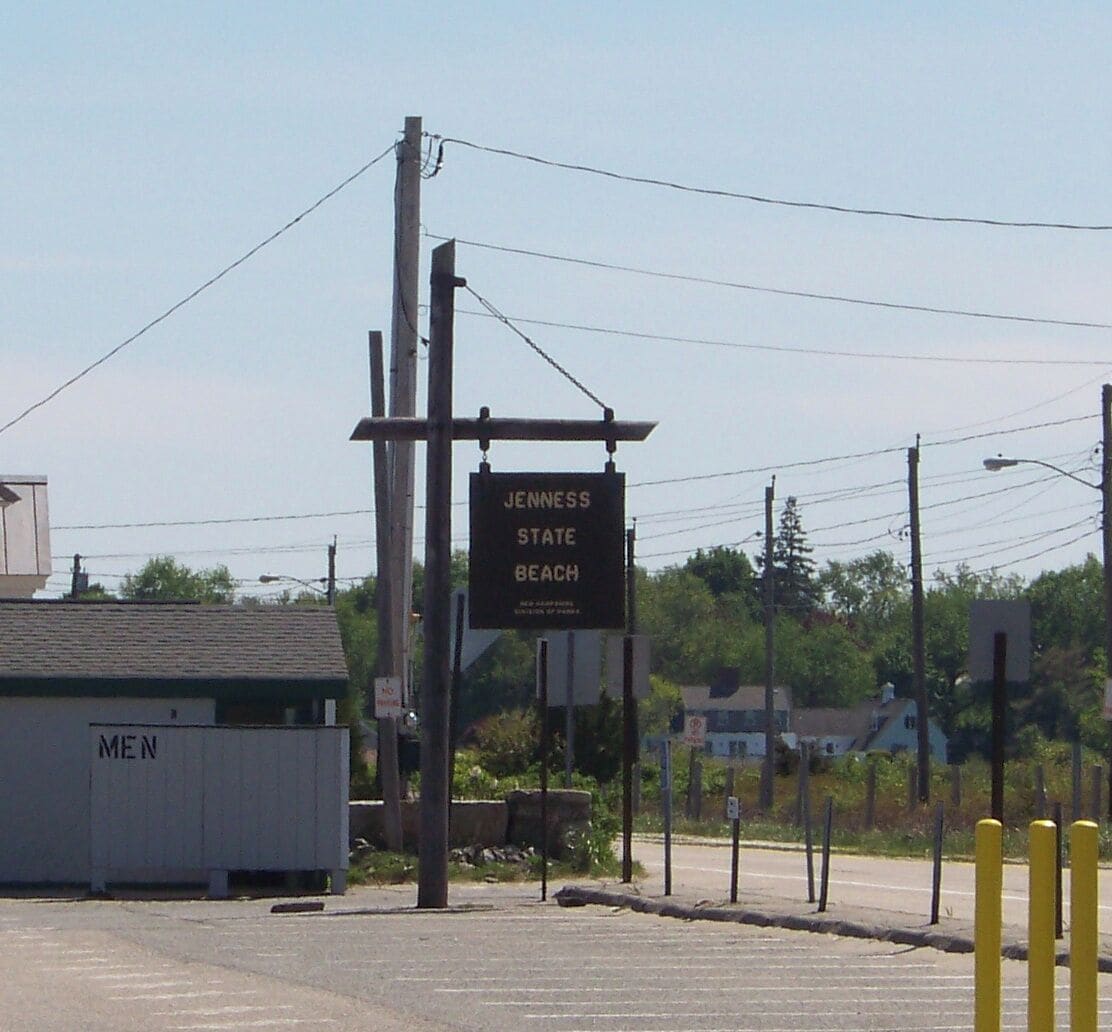 Un panneau indiquant l’entrée de la plage d’État de Jenness, avec un bâtiment portant l’inscription « HOMMES » en arrière-plan.