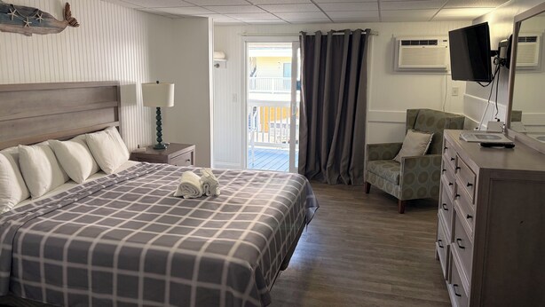 A hotel room with a bed, a chair, a dresser, a television, and a view of the outside. - Mainsail Inn & Cottages (Hampton)