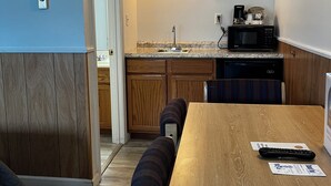 A compact kitchen with a built-in microwave, a coffee maker, and a sink. There is a wooden dining table with a bench, and a door leading to another room. - Mainsail Inn & Cottages (Hampton)