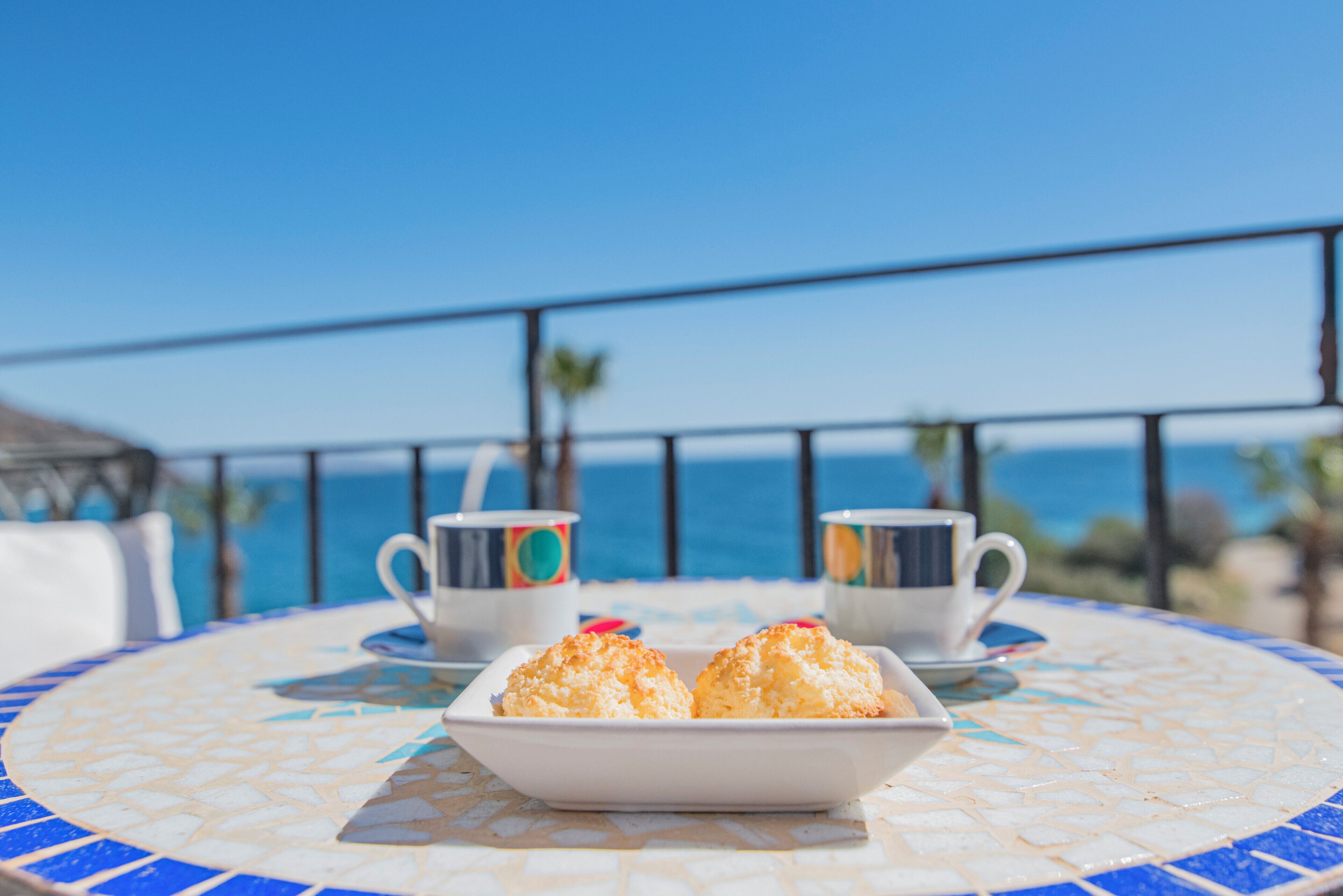 A table with a white plate of pastries and two white cups with colorful designs, set on a blue and white tiled table, overlooking a clear blue sky and ocean.