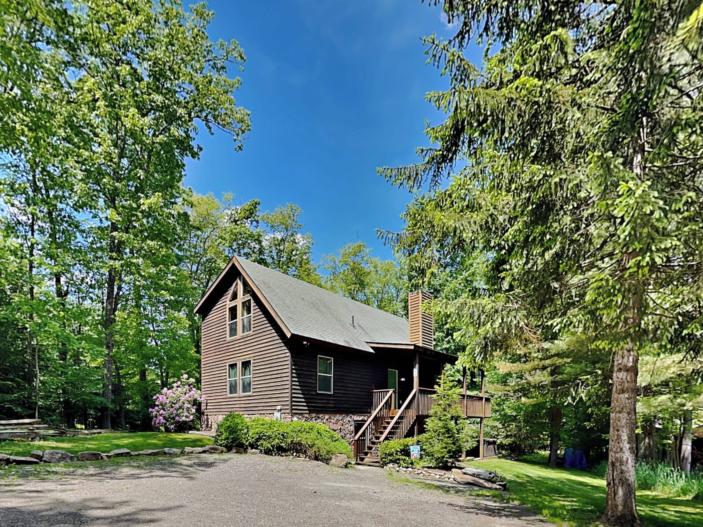 A two-story log cabin with a steeply pitched roof, surrounded by lush greenery and trees.