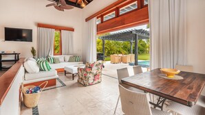 A spacious living area with a wooden ceiling, a large TV mounted on the wall, a sofa with white cushions, a floral armchair, and a dining table with chairs. - Tropical Bungalow With Pool at cap Cana A8C (Punta Cana)