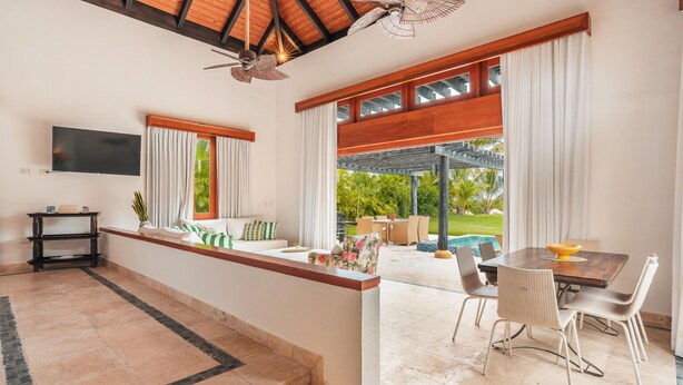 A spacious living area with a TV mounted on the wall, a dining table with chairs, and a view of a pool and greenery outside. - Tropical Bungalow With Pool at cap Cana A8C (Punta Cana)