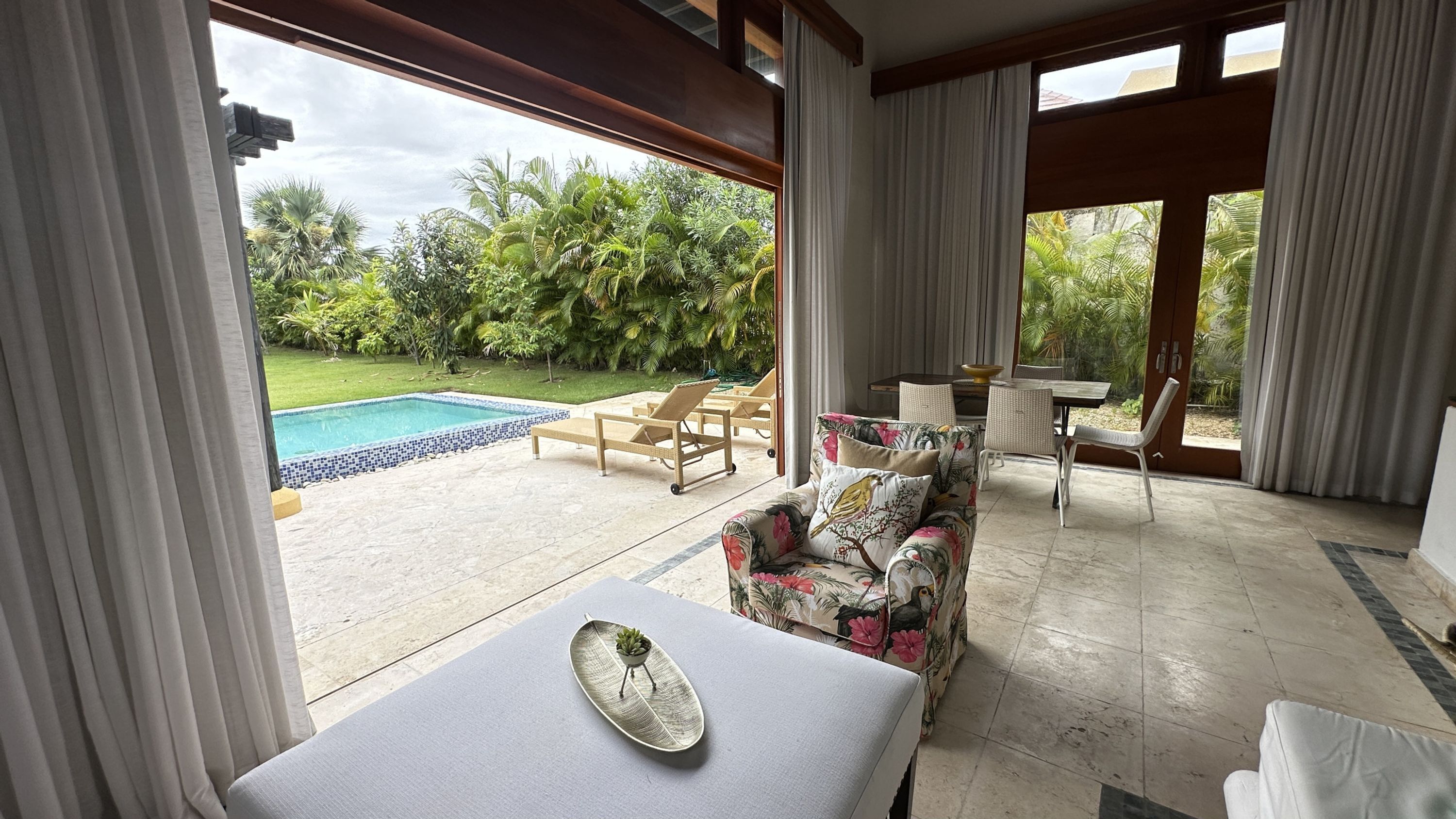 A spacious room with a view of a swimming pool and lush greenery. There is a sofa with floral cushions and a small table with a decorative plate.