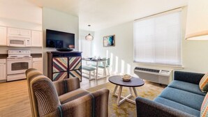 Een moderne woonkamer met een bank, fauteuils, een kleine tafel en een televisie. Er is een keukenhoek met een magnetron en een fornuis.