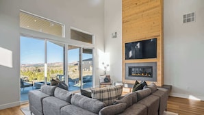A modern living room with a gray sectional sofa, a fireplace, and large windows offering a view of the outdoors. - The Meadow at Natl Parks -Hot Tub- Sleeps 10-Family (Hatch)