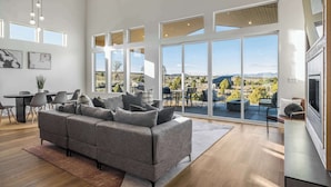 A spacious living room with a gray sofa, dining area, and large windows offering a view of the outdoors. - The Meadow at Natl Parks -Hot Tub- Sleeps 10-Family (Hatch)