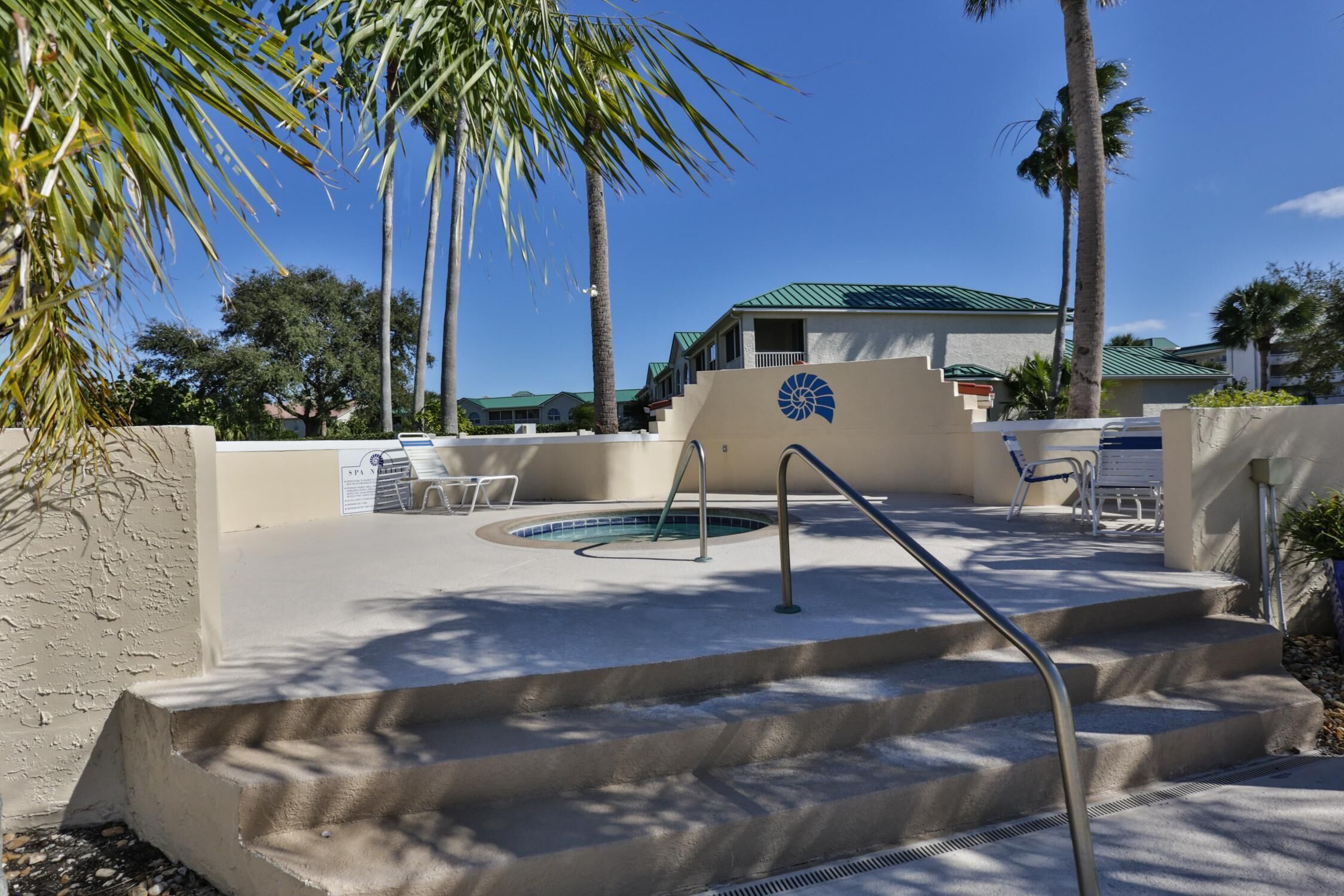 A pool area with steps leading to a lounge area, surrounded by palm trees and a building with a green roof.