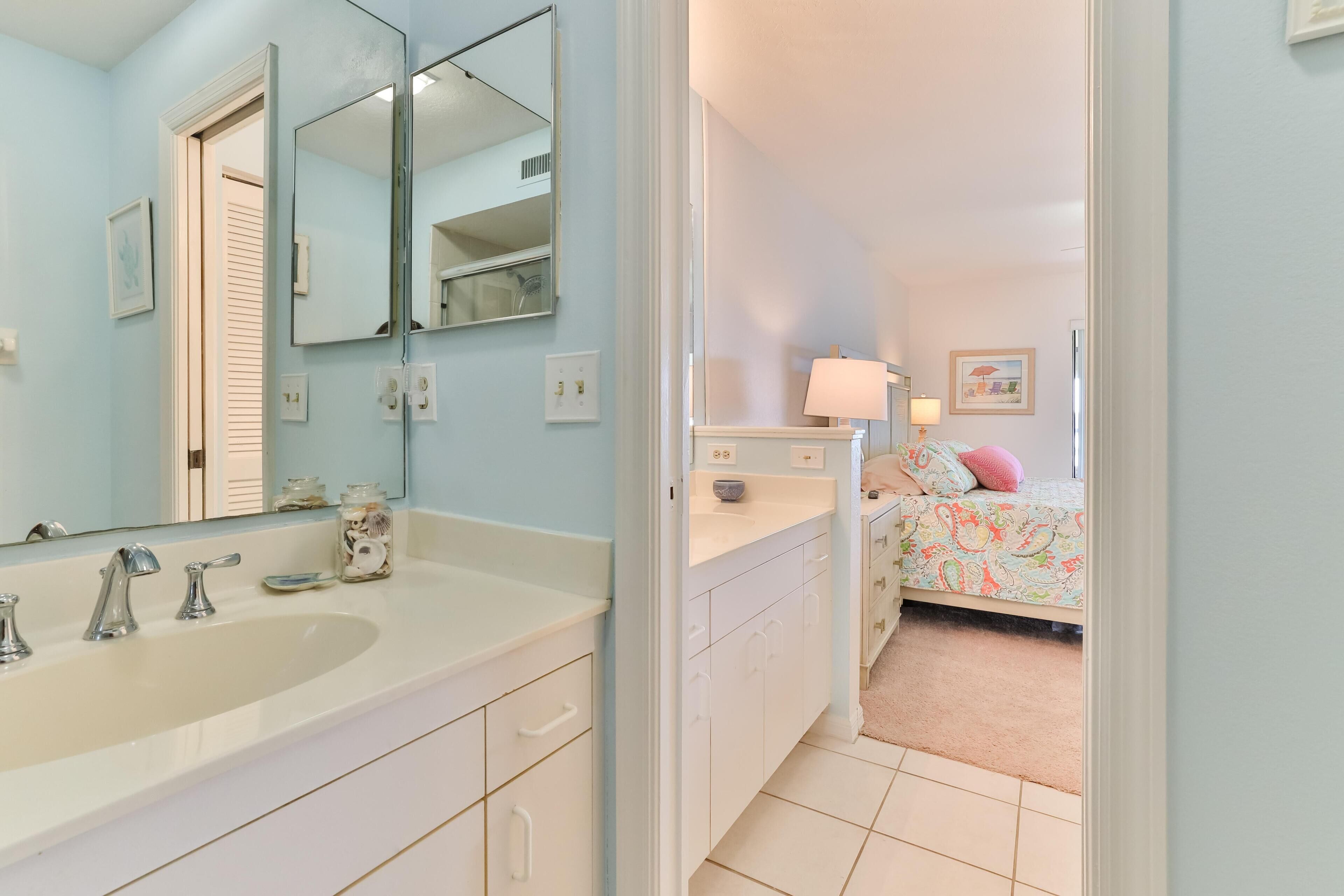 A bathroom with a double sink, a large mirror, and a door leading to a bedroom with a bed and a nightstand.