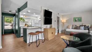 Un appartement moderne comprenant une cuisine, un coin repas et un salon. Il est équipé d’un grand téléviseur à écran plat, d’un canapé et d’une table basse en verre.