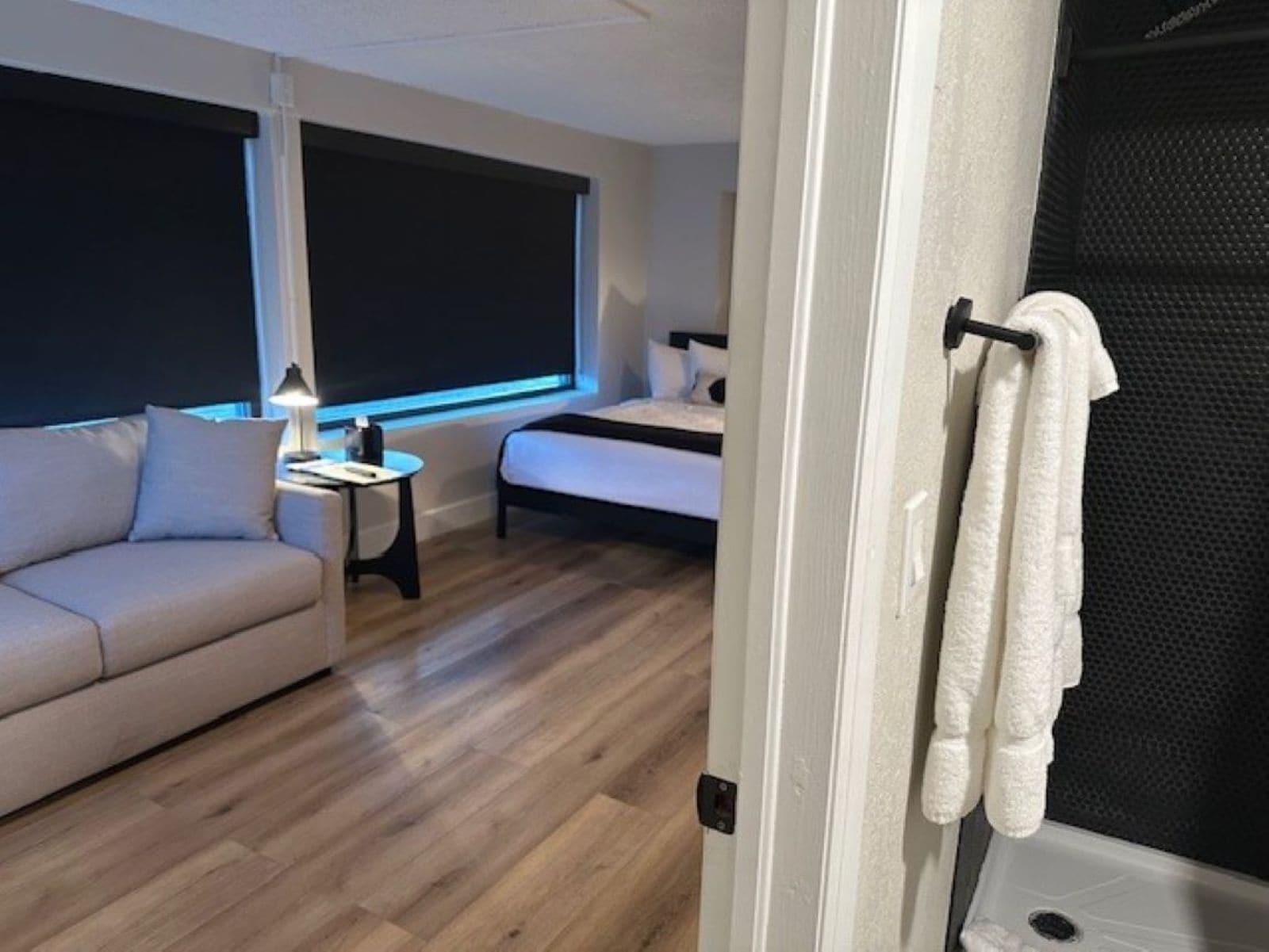 A modern bedroom with a bed, a sofa, a small table with a lamp, and a window with blinds.