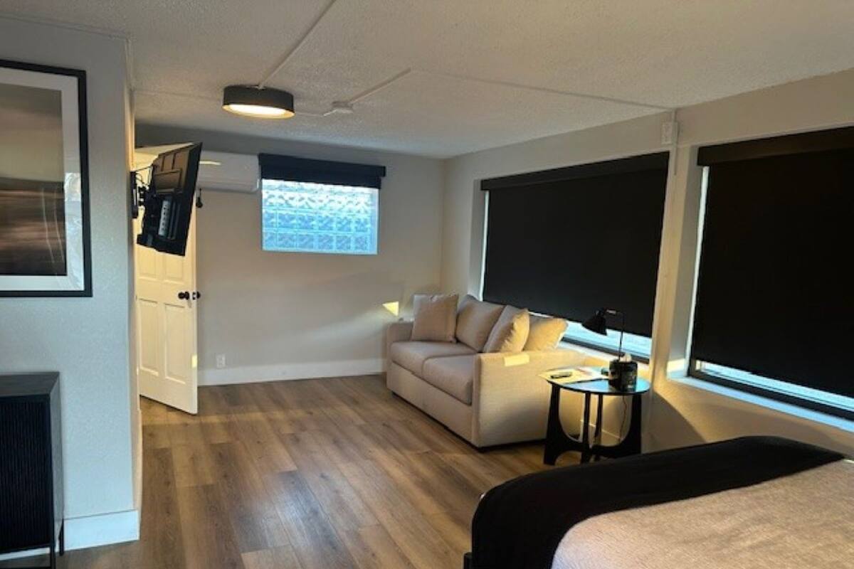 A modern hotel room with a bed, a sofa, a small table, a TV, and a window with blinds.