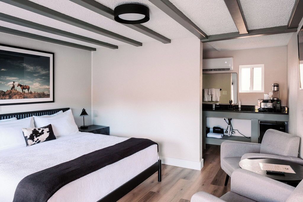 A modern hotel room with a large bed, a desk, a chair, and a kitchenette.