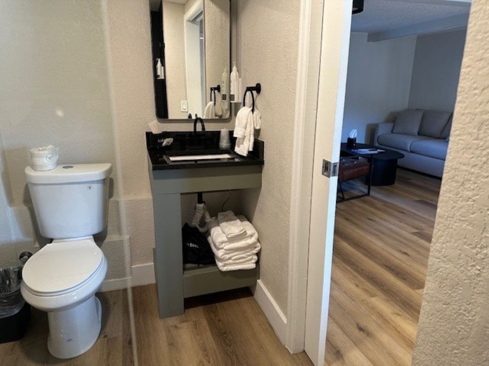 Une petite salle de bain comprenant des toilettes, un lavabo et un miroir. On peut voir un porte-serviettes, et le sol est recouvert de parquet.